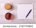 Small photo of a note book with empty space for writing, a pen and a tea cup with black tea and a plete with cookies. minimalism style. top view