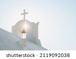 White Orthodox Church belfry with cross and bell in sunshine on sky background. Santorini island, Greece
