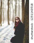 Small photo of Portrait of young womqn in stylish coat and sunglasses standing near the tree tunk in the snowy park on sunny winter day