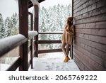 Charming young woman standing on balcony of modern wooden cottage and enjoying view on snowy forest. Beautiful winter weather. Travelling concept.