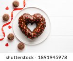 Chocolate heart shaped bundt cake covered by chocolate glaze and red sprinkles and chocolate truffles. Saint Valentine's day dessert. Festive food. Top view. Copy space. White wooden background.