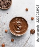 Small photo of Chocolate cream spread. Sweetened cocoa paste in ceramic bowl, teaspoon, milk chocolate chips, hazelnuts. Gray concrete background. Top view. Delicious breakfast ingredient, dessert or snack.