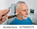 Small photo of Audiologist presents BTE hearing aid for mature hearing impaired man to treat his deafness, close-up. Audiology, auditory solutions