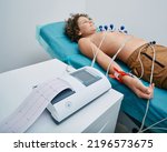 Small photo of Cardiograph with ECG printout for teenager boy patient lying in medical bed with vacuum sensors. Heart electrocardiography for children