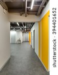 Small photo of Corridor with white walls. Right wall is with orange colored zones and with passages. There are gray doors at the end of corridor. Near the nearer door there are two small tables with plants in the