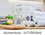 Small photo of Glass of filtered clean water with reverse osmosis filter, lemons and cartridges on a table in kitchen. Concept Household filtration or purification system.