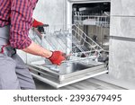 Technician or worker in uniform installs dishwasher into the kitchen furniture. Repairman wear worker suit repairing maintenance of dishwasher. Master in protective gloves fix dishwasher.