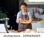 Portrait of an active elderly woman cooking in the kitchen. Grandma has prepared delicious pastries and is holding a plate of whites. Healthy homemade food.