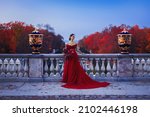 Small photo of A beautiful girl in a long red ball gown standing on the balcony against the background of an autumn park in the twilight light.
