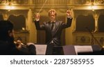 Small photo of Cinematic Close Up of Conductor Directing Symphony Orchestra with Performers Playing on Stage During Music Concert. Professional Conductor Leading Musicians Passionately in Classic Theater