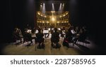 Small photo of Wide Cinematic shot of Conductor Directing Symphony Orchestra with Performers Playing Violins, Cello and Trumpet on Classic Theatre with Curtain Stage During Music Concert. Back View