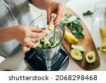 Close Up Photo of Woman Hands Making a Healthy Detox Drink in a Blender - a  Green Smoothie with Bananas, Green Spinach and Avocado. Healthy eating concept, ingredients for smoothies on the table.