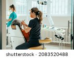 Small photo of Woman Dentist in Protective Workwear Using Dental Drill for Treating Dental Cavity on Male Patient Teeth in a Dental Clinic with Female Dental Assistant in the Background