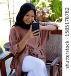 Small photo of Magelang, Indonesia. Isma is sitting while playing gadget with a smiling face in front of her house Magelang, Central Java, Indonesia on October 9, 2019.