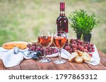 Point of view shot of two glasses of rose wine outside on a rustic wooden picnic table surrounded by red grapes, French bread, and a wine bottle.
