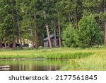 Small photo of Red Feather Lakes, Colorado, USA - July 10, 2022. Shoreline of Dowdy Lake with outdoor restroom in the trees. Green trees, tall grasses and reflective water with a building in the trees.
