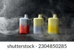 Small photo of A set of multicolored disposable electronic cigarettes on a dark background stand on a table with smoke around. The concept of modern smoking, vaping and nicotine