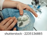 Small photo of A woman mends jeans, sews a patch on a hole, hands close-up.Mending clothes concept,reusing old jeans.