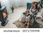 Small photo of Remote work. Young woman using laptop in a cozy armchair and plaid sitting by the fireplace with a domestic cat and drinking tea at home