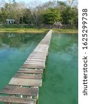 Small photo of old wooden gangplank bridge over shallow water heading towards an island with topical trees and some small buildings on.