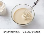Small photo of A glass bowl of dough for Finnish pancakes with a whisk and a jar of flour on a light gray background, top view. Cooking delicious homemade food