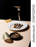 Small photo of Opened tin fish, sardine with a slice of lemon on powder colored table with black background. A fork is floating above. Abstract conception.