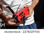 Small photo of A man holds in his hands a small red portable radio rechargeable with solar panels or manually with a crank and an included flashlight