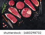 Various cuts of meat, shot from the top on a black background with salt, pepper, rosemary and knives, with copy space