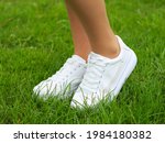 Legs of a girl in white sneakers on green grass (lawn). Close up of a pair of white shoes on the legs of a girl against a background of green grass.
