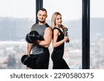 Sporty athletic young couple posing with dumbbells near window in gym.
