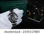 Small photo of Rolex wristwatch model cosmograph daytona oyster perpetual superlative chronometer with black ceramic bezel stainless steel body is in left hand with background green rolex package box and led light