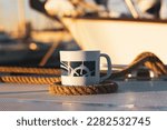 White cup with drawn helm on deck of a white yacht, ship, boat. Concept of tea coffee drinking while traveling at sea ocean. A mug among the sea ropes in morning sunshine light. Nautical wallpaper.