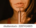Small photo of Cropped portrait of young beautiful woman, girl with sensitive lips using vibrating golden facial massager on dark black background. Preservation, prolongation of natural beauty youth Female skin care