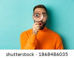 Small photo of Funny man looking through magnifying glass, searching or investigating something, standing in orange sweater against turquoise background
