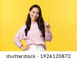 People emotions, lifestyle and fashion concept. Smiling happy good-looking asian girl giving advice, showing one finger as explain rules, make point, standing yellow background