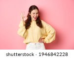 Small photo of Cheeky young woman winking, showing okay sign, give her approval, like and approve good thing. Girl make OK gesture to give permission, say yes, standing over pink background