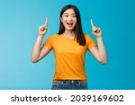 Small photo of Amused asian girl introduce new product sharing promo with you, raise hands pointing fingers up smiling broadly, look excited and upbeat, enthusiastic advertising, stand blue background