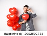 Valentines day and love concept. Sad crying man holding red heart balloon and whiping tears, standing single and miserable, being heartbroken, white background