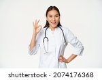 Small photo of Confident female doctor, asian physician in medical uniform and stethoscope, showing okay sign and nod pleased, praising, recommending smth good, white background