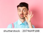 Small photo of Close up of funny caucasian guy touching his french moustache, pucker lips and looking silly, standing over pink background