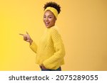 Small photo of Sassy cute feminine african-american girl 20s in headband sweater smiling cheeky flirty look camera turning standing profile pointing left introdue awesome new product showing way, yellow background