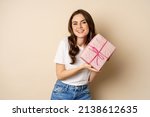Celebration and holidays concept. Happy young woman holding gift wrapped in pink box, receive present, looking amazed and surprised, standing over beige background