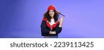 Small photo of Stylish and snobbish arrogant curly-haired woman in warm beanie rolling curl on finger smirking and looking with contempt at camera, scorning person as being too cool over blue background.