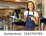 Smiling asian barista girl, wears apron, shows credit card machine for processing payment, suggest to pay contactless, standing in coffee shop.