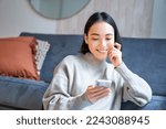 Portrait of smiling asian woman looking intrigued at smartphone screen, interested with smth on mobile phone, sitting coy on floor in living room.