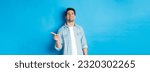 Small photo of Confident handsome guy smiling sassy, pointing finger right at copy space, showing advertisement, standing over blue background.