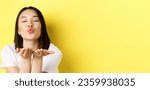 Small photo of Valentines day concept. Close up of beautiful asian woman pucker lips and holding hands near mouth, blow air kiss at camera with closed eyes, standing over yellow background.
