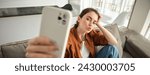 Small photo of Portrait of cute young woman, modern girl taking selfies on smartphone app, posing on couch, extends her white mobile phone to take picture, pucker lips for photo.