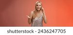 Small photo of Joyful talkative outgoing attractive blond woman talking friend smartphone gesturing amused smiling broadly retelling fresh rumors after party wearing silver stylish dress, red background.