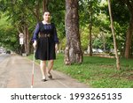 Small photo of Asian blind person woman walking on sidewalk with a long white cane a mobility tool used to detect objects in the path, also helpful for onlookers in identifying the user as blind or vision disability